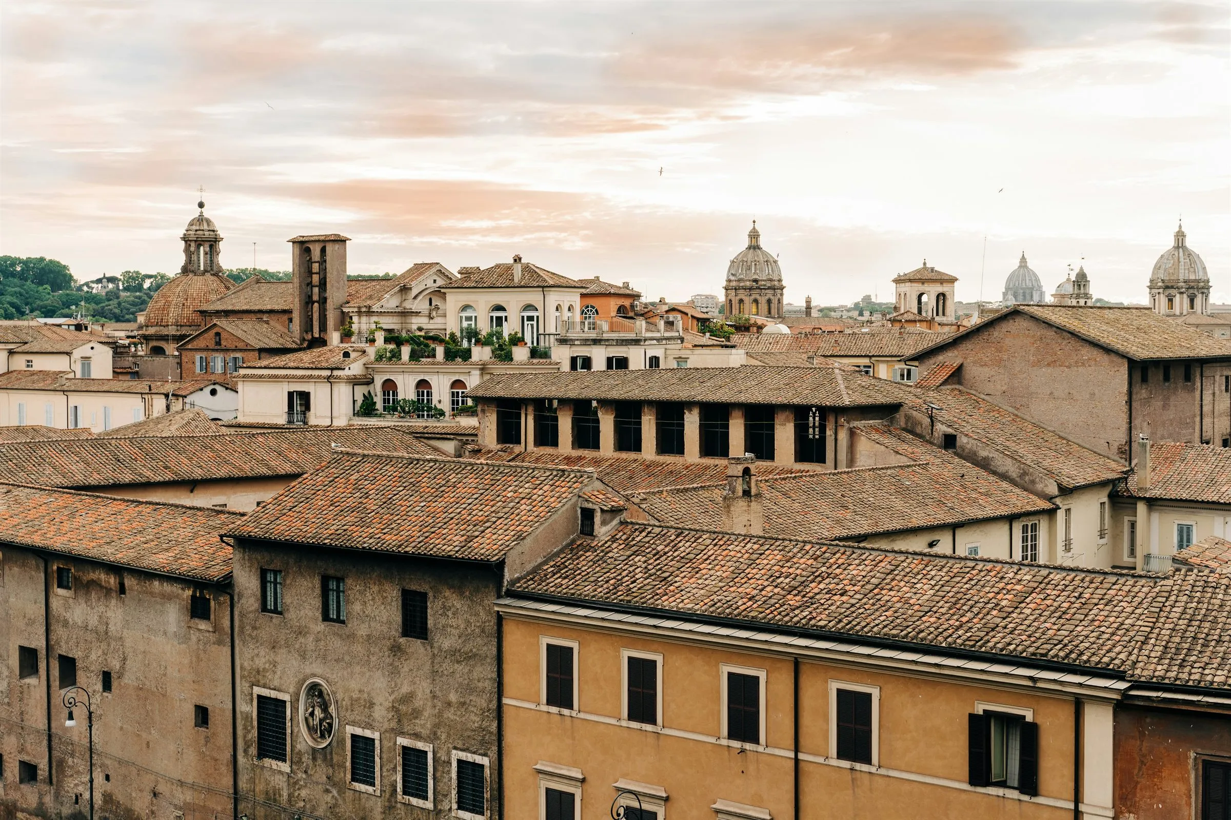 Terracotta rooftops of central Rome with church domes along the skyline under a soft, pale evening sky.